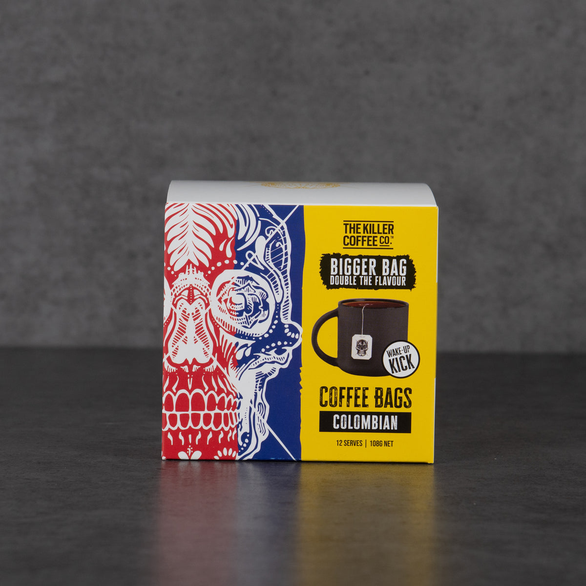 A boxed coffee bag product standing upright on a dark surface against a gray wall. The package is mostly white with a bright yellow front panel and a red-and-blue illustrated side panel.